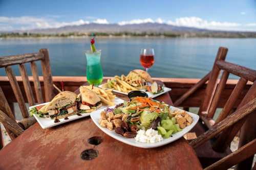 A scenic table by the water featuring burgers, fries, a colorful salad, and two vibrant drinks.