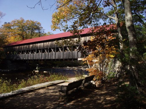 A wooden covered bridge with a red roof spans a river, surrounded by autumn foliage and a nearby bench.