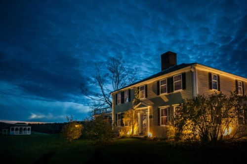 A charming house illuminated at night, surrounded by trees and a dramatic sky filled with clouds.