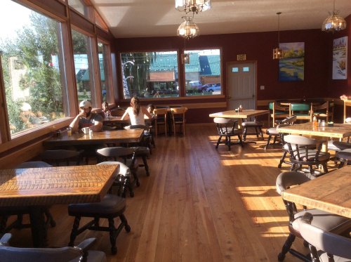 Brightly lit restaurant interior with wooden tables, chairs, and large windows showing a view outside. Two people are seated.