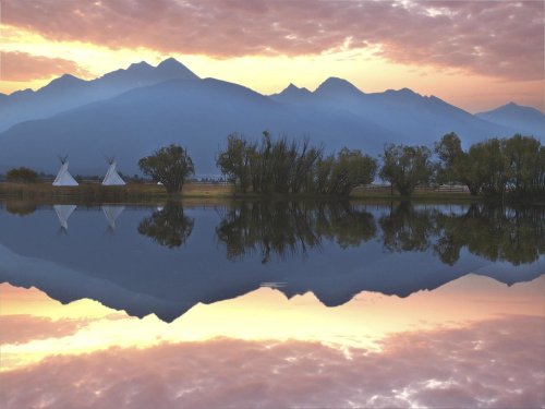 Sunset over mountains reflected in calm water, with teepees and trees along the shore.