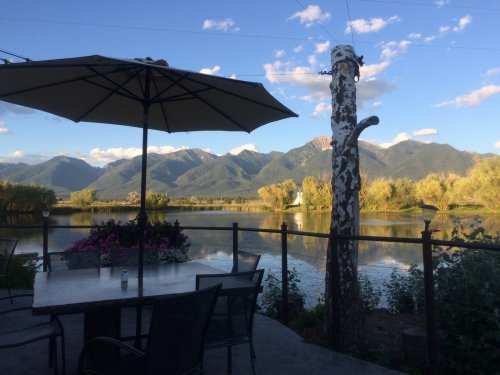 A serene lakeside view with mountains in the background, featuring a table under an umbrella and lush greenery.
