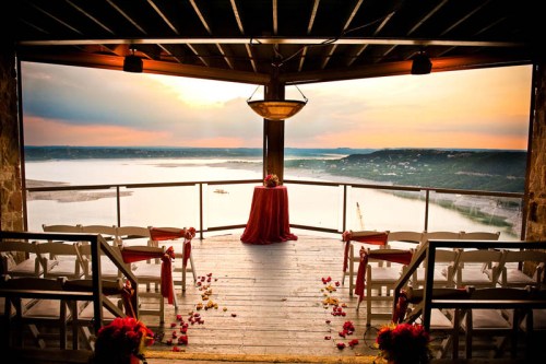 A scenic wedding venue with chairs arranged, a table at the center, and a beautiful sunset over the water in the background.