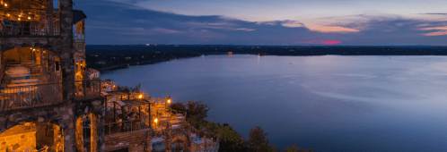 A scenic view of a lake at dusk, with illuminated terraces and a colorful sky reflecting on the water.