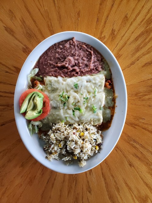A plate of enchiladas topped with cheese, served with rice, refried beans, lettuce, tomato, and avocado.