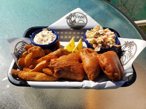 A basket of fried fish, potato wedges, coleslaw, and a creamy dip, garnished with lemon wedges.