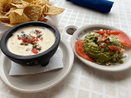 A bowl of creamy queso with diced tomatoes and a plate of guacamole topped with tomatoes, served with tortilla chips.