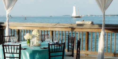 A beautifully set table with white flowers overlooks a calm sea and a sailing boat in the distance.