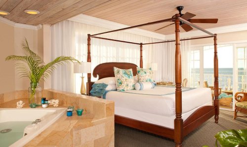 Bright, airy bedroom with a four-poster bed, tropical decor, and a view of the ocean through large windows.
