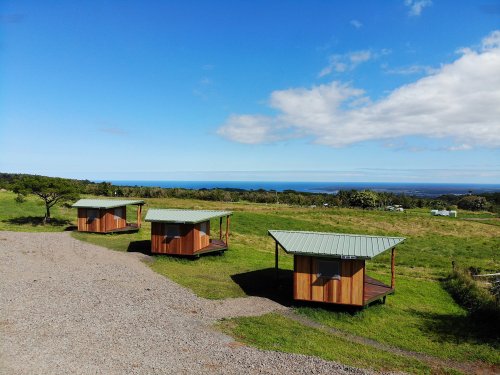 Three wooden cabins with green roofs sit on a grassy hillside overlooking the ocean under a blue sky.