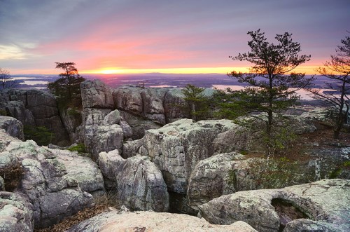 Sunset over rocky terrain with trees, casting vibrant colors across the sky and illuminating the landscape.