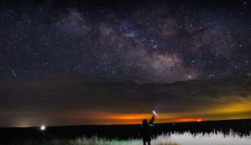 A person stands in a field at night, pointing a light towards a starry sky filled with the Milky Way.