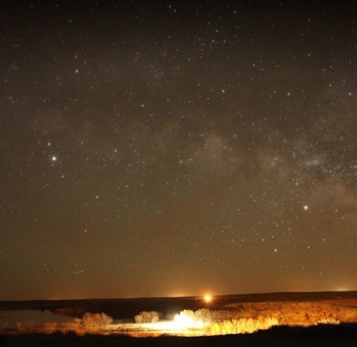 A starry night sky over a dark landscape, with a faint glow from distant lights and a hint of the Milky Way.
