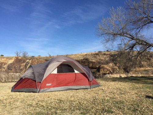 A red and gray tent set up on grassy ground with a rocky hillside and clear blue sky in the background.