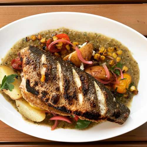 Grilled fish fillet on a bed of sauce, garnished with corn, onions, and herbs, served on a white plate.