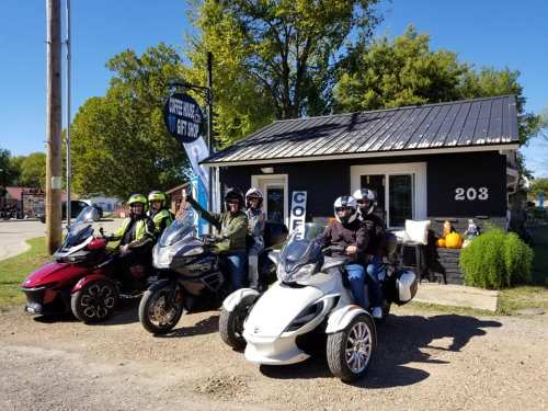 A group of motorcyclists poses with three trikes in front of a small building, surrounded by trees and autumn decor.