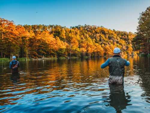 Two anglers fishing in a serene lake surrounded by vibrant autumn foliage and calm waters.