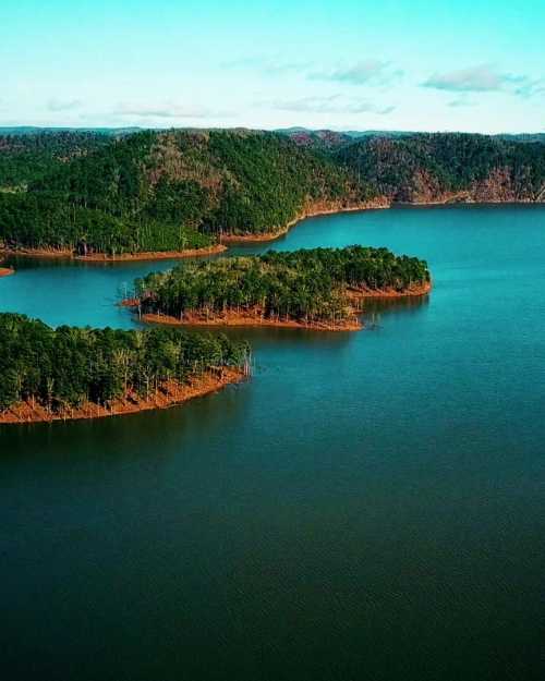 Aerial view of a serene lake surrounded by lush green islands and forested hills under a clear blue sky.