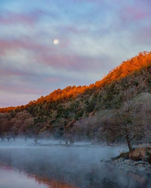 A serene landscape at dawn, featuring a misty river, illuminated hills, and a full moon in a colorful sky.