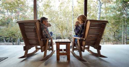 A couple sits in rocking chairs on a porch, enjoying drinks and the view of trees in autumn.