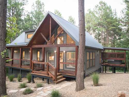 A modern cabin with a sloped roof, large windows, and a wooden deck, surrounded by trees and gravel landscaping.