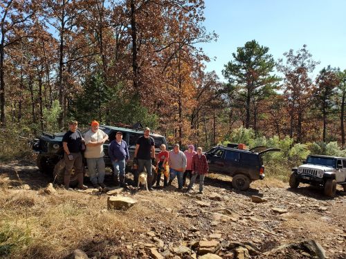A group of people stands on a rocky trail in a forest, with off-road vehicles parked nearby and autumn foliage in the background.