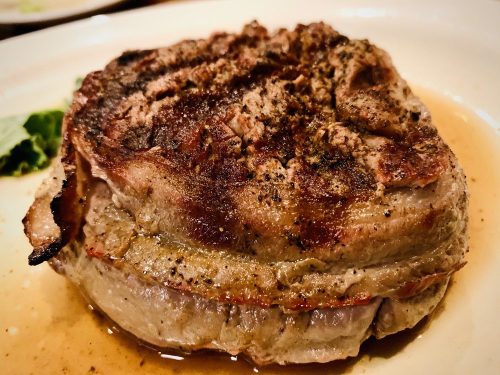 A perfectly cooked, grilled steak with a seared crust, resting on a plate with a light sauce and garnish.