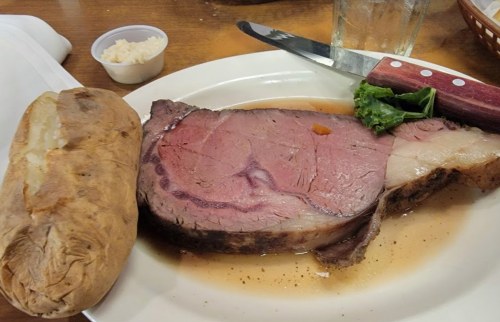 A plate featuring a large slice of prime rib, a baked potato, and a small cup of horseradish on the side.