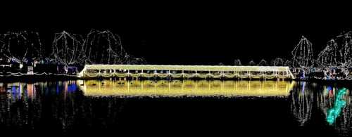 A beautifully lit bridge reflects in a dark lake, surrounded by glowing trees and festive decorations.