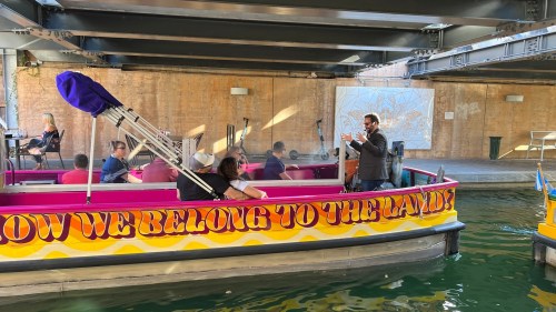 A colorful boat with people onboard, under a bridge, with a speaker addressing the group. Art displayed on the wall.