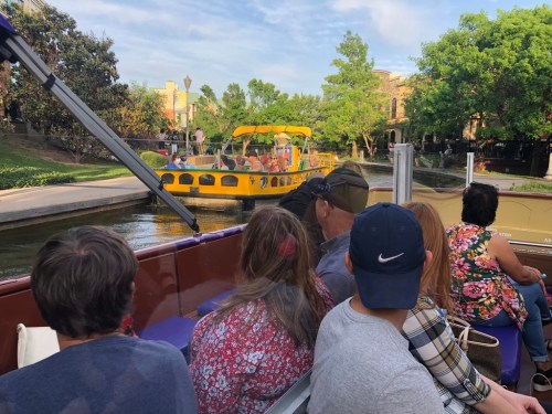 A group of people on a boat enjoying a scenic ride along a canal with greenery and another boat in view.