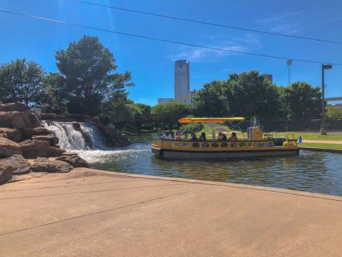A yellow boat with passengers floats on a calm lake near a waterfall and green trees under a clear blue sky.