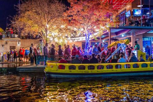 A vibrant night scene with a yellow boat and a crowd gathered by the water, illuminated by colorful lights and autumn trees.