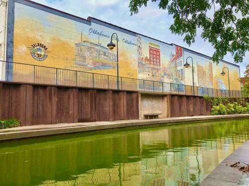 Mural depicting Oklahoma landmarks on a wall beside a green canal, with trees framing the scene.