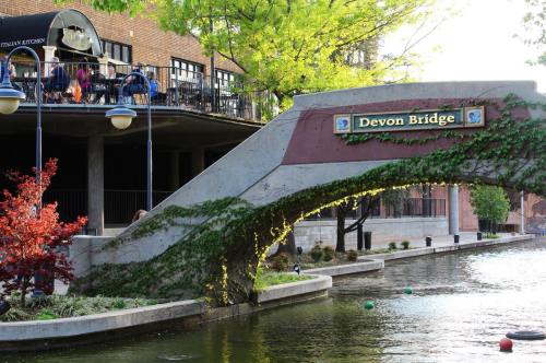 A stone bridge with "Devon Bridge" sign, surrounded by greenery and a canal, with people on a nearby balcony.
