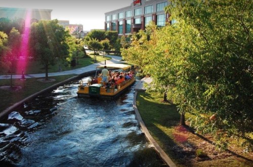 A boat with passengers glides through a scenic waterway lined with trees and greenery, near a building in the background.