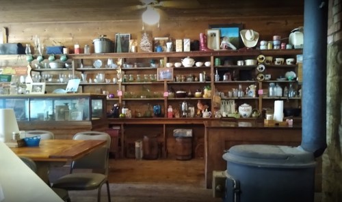 A rustic interior featuring a wooden shelf filled with various jars, dishes, and vintage items, alongside a cozy dining area.