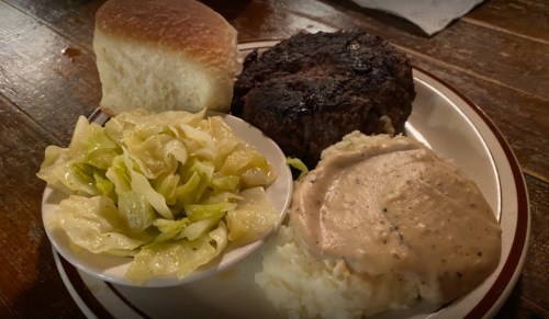 A plate with a hamburger patty, mashed potatoes with gravy, sautéed cabbage, and a roll.