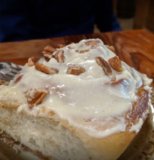 Close-up of a cinnamon roll topped with cream cheese frosting and chopped pecans on a wooden table.