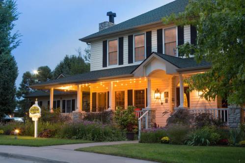A charming two-story house with a porch, illuminated by string lights, surrounded by lush greenery and colorful flowers.