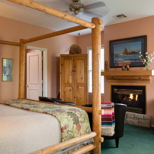 Cozy bedroom with a wooden bed frame, fireplace, and colorful decor, featuring a chair and natural light from windows.