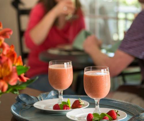 Two glasses of strawberry smoothie on a tray, with fresh strawberries and flowers in the foreground, and people in the background.