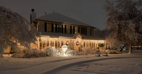 A cozy house adorned with holiday lights, surrounded by snow-covered trees and a winter landscape at night.