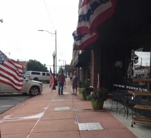 Two people walk along a sidewalk lined with American flags and planters in a small town setting.