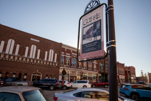 A street sign in a historic downtown area, with buildings and parked cars in the background under a clear sky.