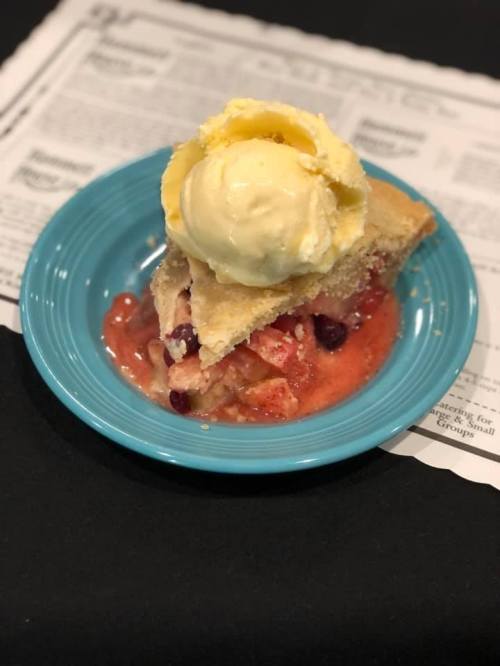 A slice of fruit pie topped with a scoop of vanilla ice cream on a blue plate, resting on a newspaper.