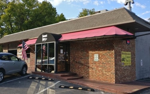 A brick building with a pink awning, featuring large windows and American flags outside. Parking area visible.