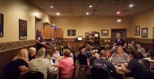 A busy restaurant scene with diners at tables and servers attending to customers in a cozy, warmly lit setting.