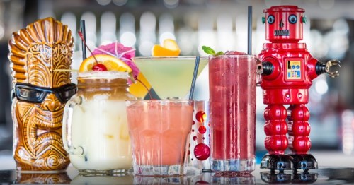 A colorful display of five cocktails, including a tiki mug, a robot figure, and garnishes like fruit and umbrellas.