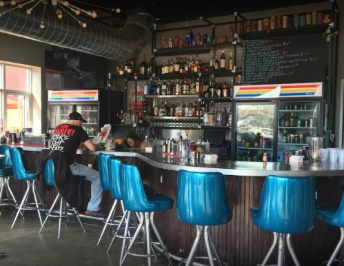 A modern bar with blue stools, shelves of liquor, and a person sitting at the counter reading a menu.